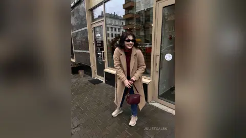 Smiling woman in a beige wool coat and burgundy sweater walking on a city sidewalk near a storefront.