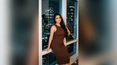 Smiling woman in a brown ribbed midi dress standing by a window with a night city skyline view.