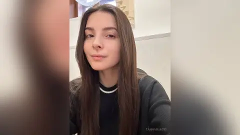 Young woman with long brown hair wearing a black sweater in a bright indoor setting with a calm expression.