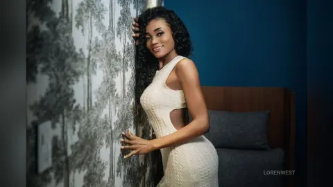 Smiling woman in a white knit dress leaning against patterned wallpaper in a moody, elegant room.