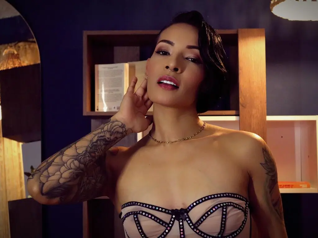 Tattooed woman in a beige lace corset posing confidently in a warm, moody indoor setting.