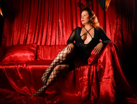 Woman in black lace bodysuit and fishnet tights sitting on a red satin sofa against dramatic red curtains.