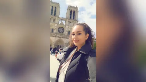 Smiling woman in a black puffer jacket taking a selfie in front of the Notre Dame Cathedral in Paris.