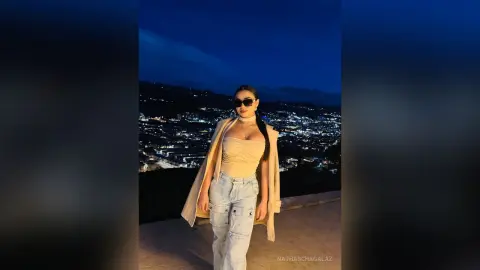 Stylish woman in a beige halter top and light jeans posing against a glowing city skyline at night.