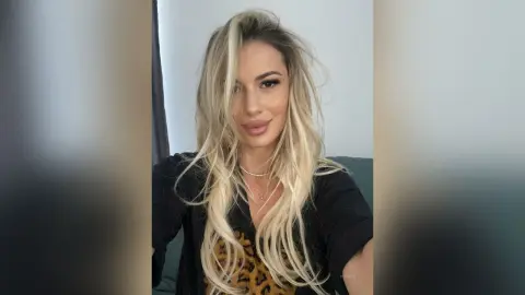 Blonde woman in a black cardigan and leopard print top smiling in a cozy indoor setting.
