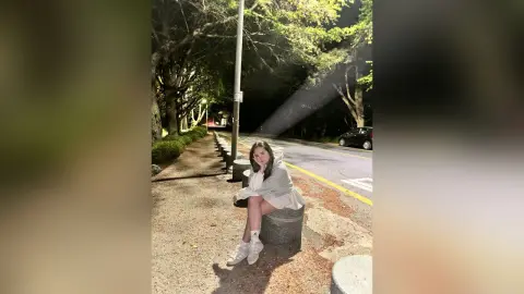 Young woman in a white sweatshirt and shorts sitting on a concrete bollard on a quiet street at night.