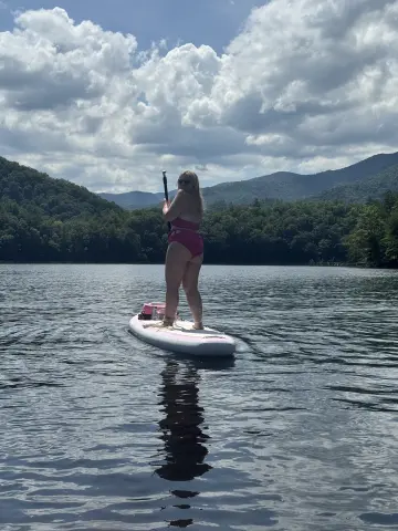 Woman in pink bikini paddleboarding on a calm lake surrounded by lush green mountains under a cloudy sky.