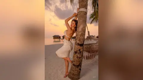 Elie poses naked against a palm tree on a beach at sunset, wearing a white string bikini top and skirt that accentuates her curves.