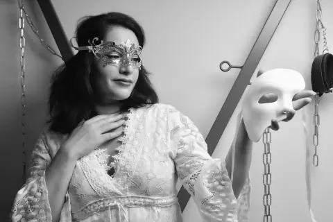 Elegant woman in white lace gown holding a masquerade mask in a moody black and white studio setting.