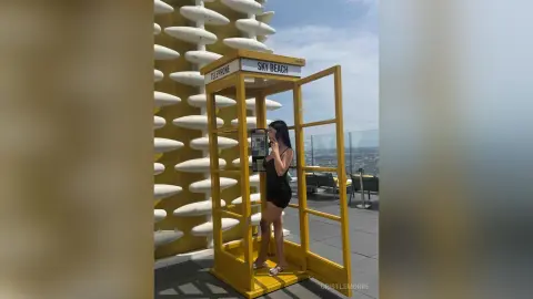 Stylish woman in a black dress using a vintage yellow telephone booth at a sunny rooftop Sky Beach lounge.
