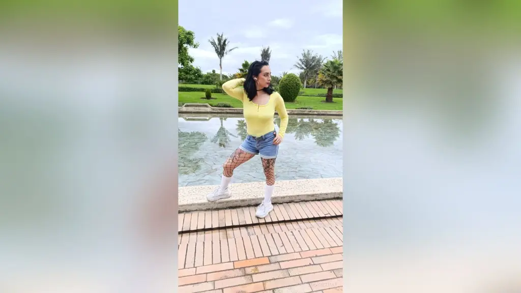 KandyPariston — Young woman in a yellow long-sleeve top and denim shorts posing by a garden pond on a sunny day.