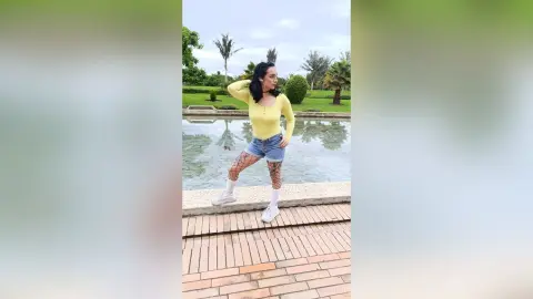 Young woman in a yellow long-sleeve top and denim shorts posing by a garden pond on a sunny day.