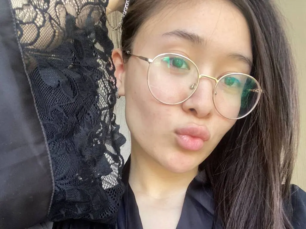 Young woman with round glasses and black lace top posing for a casual selfie in soft natural light.