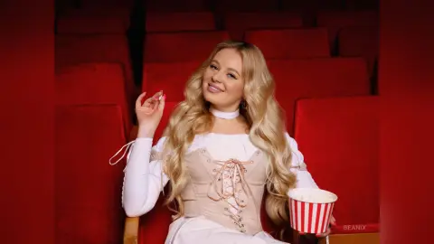 BeaNix smiles while sitting in red theater seats wearing a white lace-up corset top and white choker.