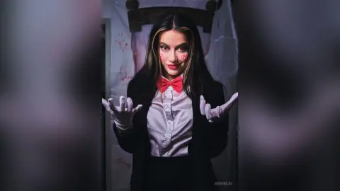 JessieLiu poses in a white button-down shirt, red bow tie, and black blazer with white gloves, featuring clown-style face paint.