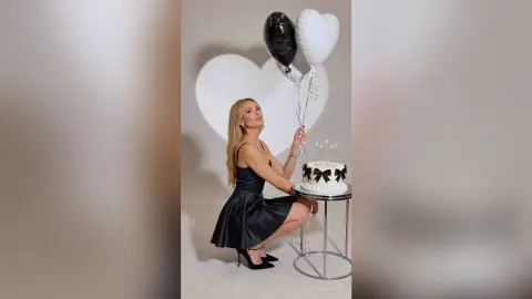 SerenaStar poses in a black satin dress next to a white heart backdrop, holding black and white balloons and a small cake.