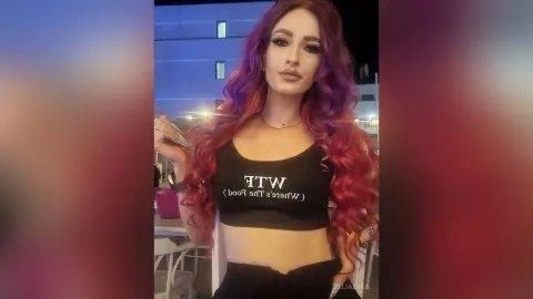 Woman with long wavy red hair wearing a black WTF crop top in a nighttime outdoor setting.