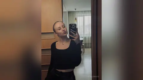 Young woman in a black long-sleeve top taking a moody mirror selfie in a modern minimalist room.