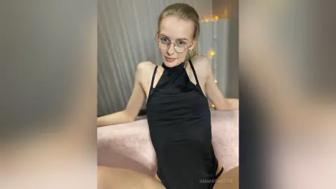 Blonde woman in a black halter neck top and glasses posing in a cozy, dimly lit room.