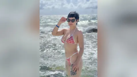 Young woman in a red checkered bikini and sunglasses standing on a sunny beach by the ocean waves.