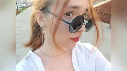Redheaded woman in a white shirt and round sunglasses smiling outdoors in bright daylight.