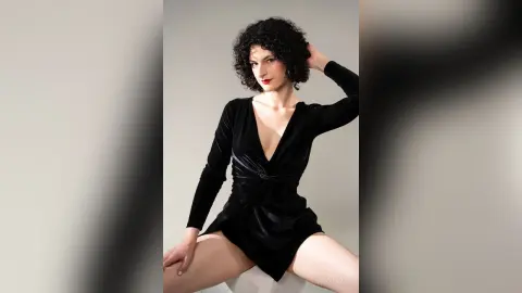 RoxyGraff poses in a black velvet long-sleeve wrap dress with a deep v-neck, showing her legs against a plain grey background.