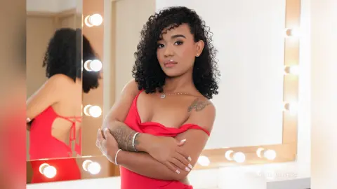 Young woman with curly hair in a red dress posing in front of a lighted vanity mirror.