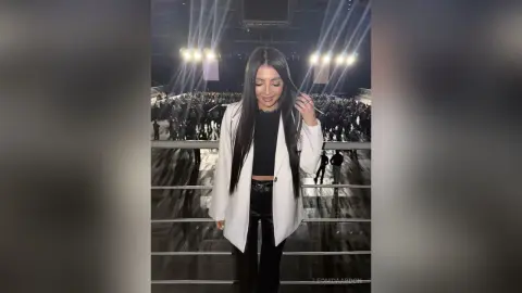 LeonidaAbdon poses in a white blazer and black top with long, straight dark hair, standing overlooking a large, dark crowd or stage area.