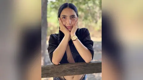Smiling woman in a black shirt leaning on a wooden fence in a sunny outdoor forest setting.