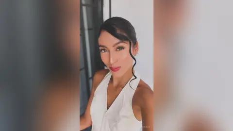 LaraOlson poses in a white sleeveless top with her dark hair slicked back and a single curl framing her face against a plain white wall.