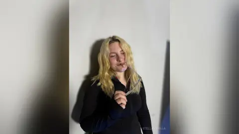 StellaVanilla with long blonde hair, wearing a black long-sleeved top and holding glasses up to her face against a plain white wall.