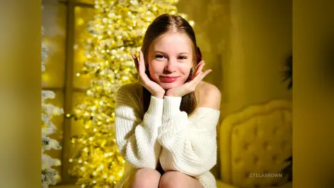 StelaBrown smiles while resting her chin on her hands, wearing a white knit sweater against a glowing yellow Christmas tree.