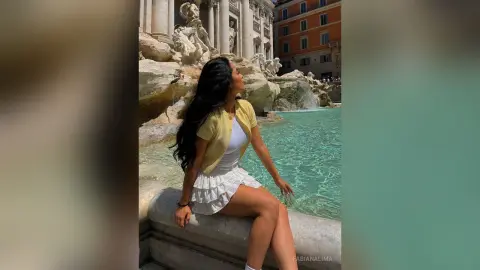 FabianaLima sits on the edge of a stone fountain wearing a white skirt and a yellow cardigan.