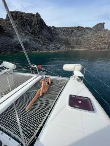 Relaxed woman in a black bikini lounging on a catamaran net in a scenic rocky cove.