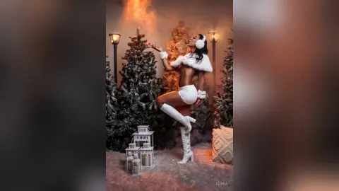 Glamorous woman in white winter outfit posing in a snowy forest setting with warm festive lighting.