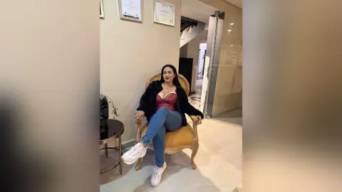 Stylish woman in a black jacket and red corset top sitting on a gold chair in a modern minimalist room.