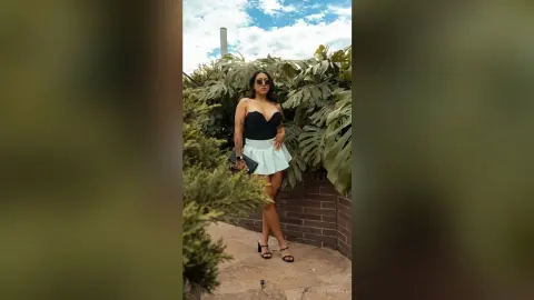 Stylish woman in a black corset and light blue mini skirt posing outdoors amidst lush tropical greenery.