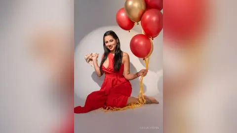 Smiling woman in a red jumpsuit celebrating with red and gold balloons in a festive studio setting.