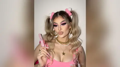 Glamorous woman with pink pigtails and dramatic eye makeup wearing gold jewelry and a pink top.