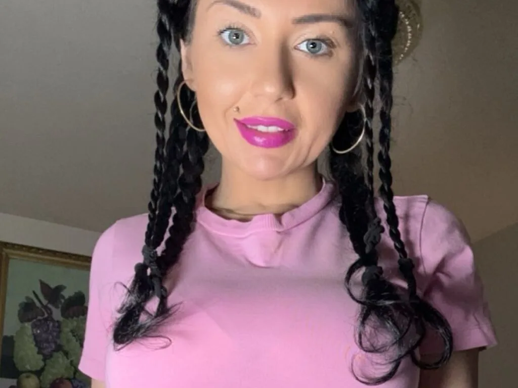 EdaVanesa with long black hair and a nose piercing looks into the camera while wearing a white tank top.