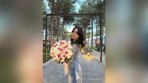 Smiling woman in a grey sweater holding a large bouquet of pink roses in a peaceful outdoor garden setting.
