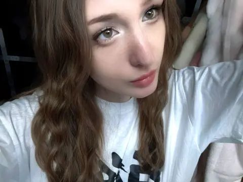 Young woman with wavy brown hair wearing a white t-shirt in a cozy indoor setting.