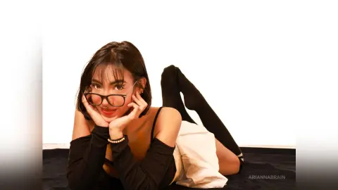 Young woman with glasses in a black top and white skirt relaxing on a dark floor against a white background.