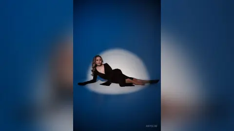 Elegant woman in a black long-sleeve dress reclining in a dramatic blue spotlight studio setting.