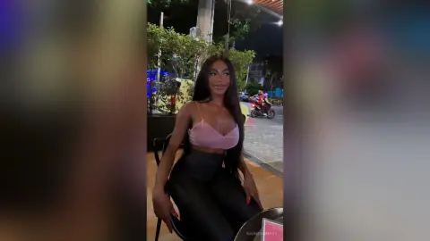 Glamorous woman in a pink crop top and black pants sitting at an outdoor night cafe.