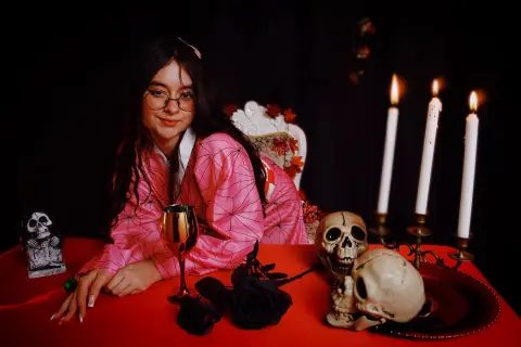 Young woman in pink kimono sitting at a spooky Halloween table with skulls, candles, and black roses.