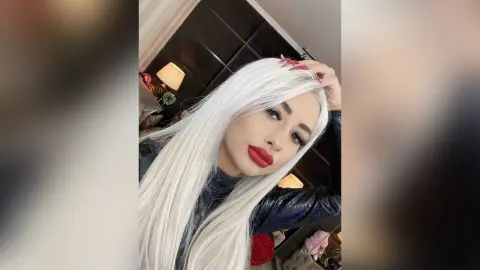 Glamorous woman with long platinum blonde hair and bold red lipstick posing in a dark room.