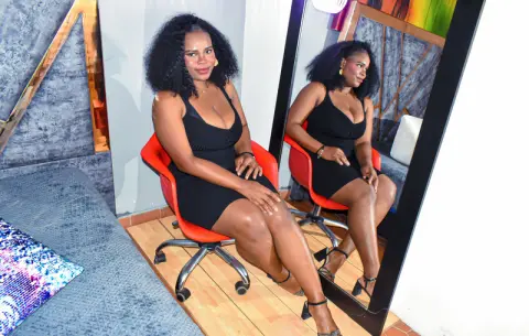 SkyllerFoxy — Confident woman in a black cocktail dress sitting in an office chair reflecting in a mirror.