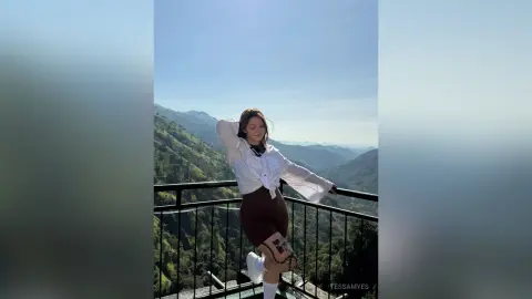 Young woman in white blouse and brown shorts posing on a scenic mountain balcony under a clear blue sky.