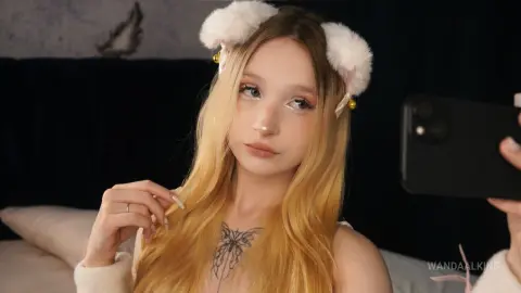 Blonde woman in white fuzzy cat ears taking a selfie in a cozy, dark bedroom setting.